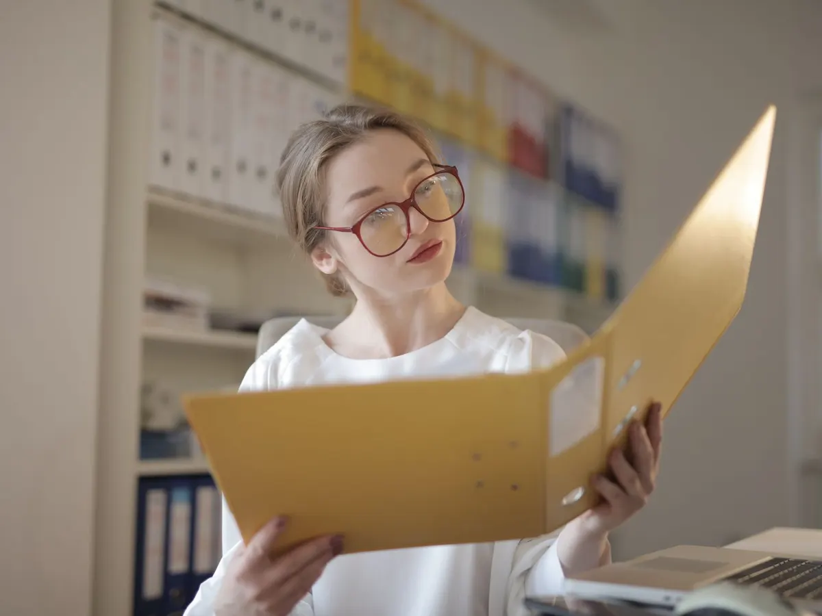 Una mujer con lentes revisa el contenido de una carpeta en una oficina con archiveros