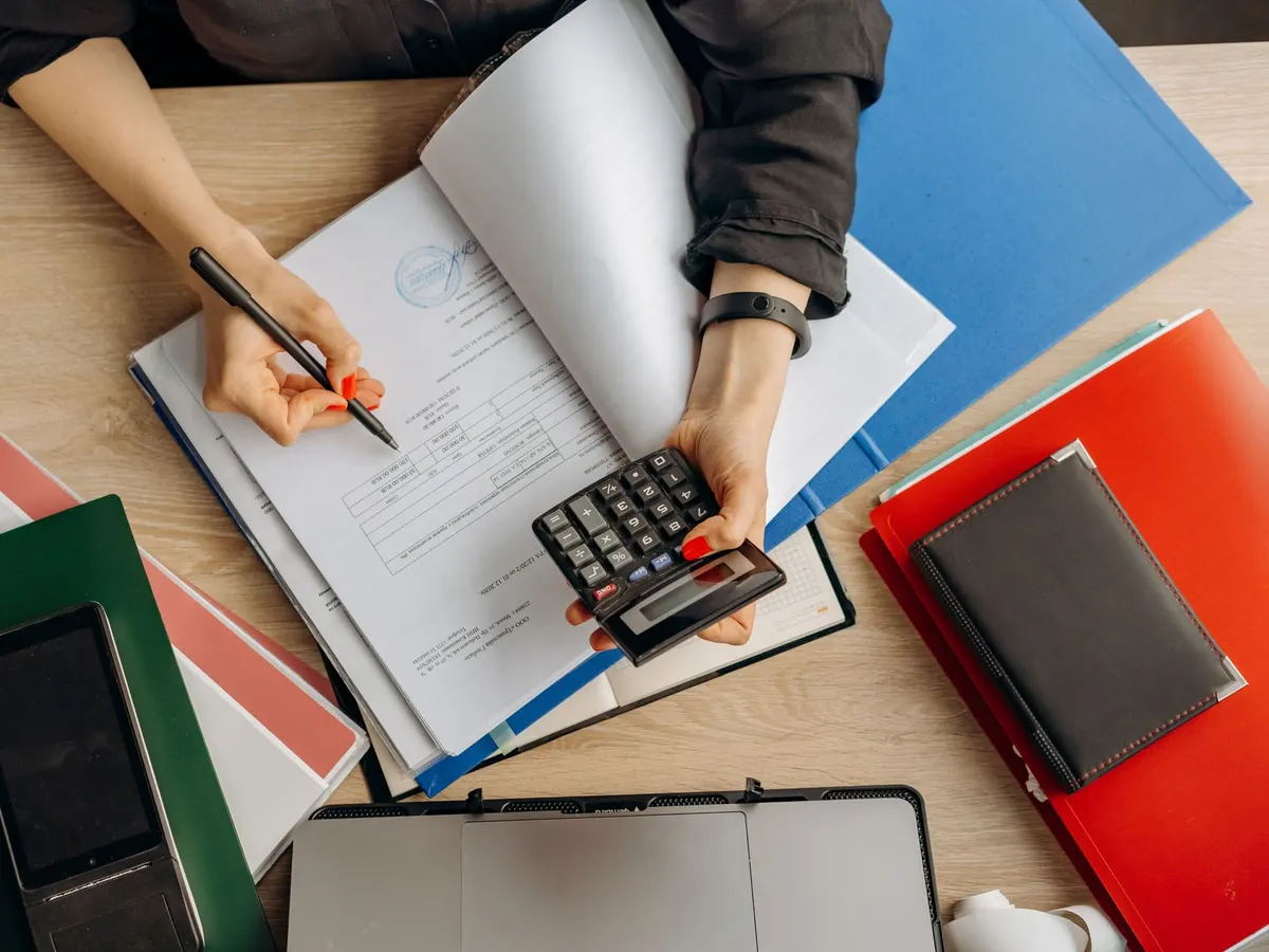 Vista cenital de una persona que anota cifras en un cuaderno con una calculadora, entre carpetas de colores