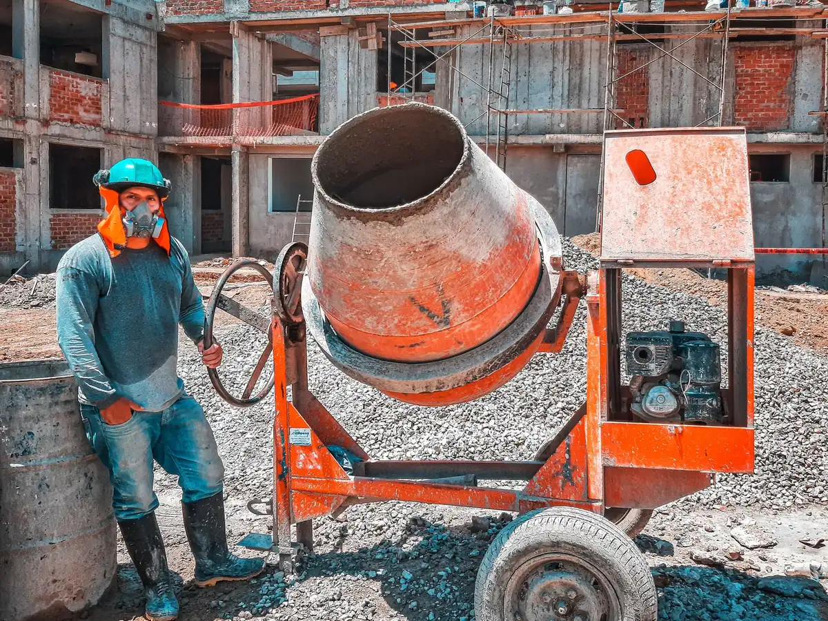 Trabajador con casco y mascarilla junto a una revolvedora de concreto en una obra en construcción
